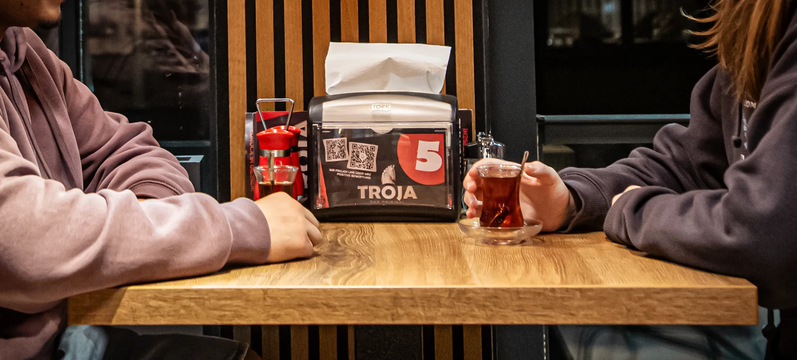 Zwei Gäste an einem Holztisch bei Troja, eine Person hält ein Teeglas.
